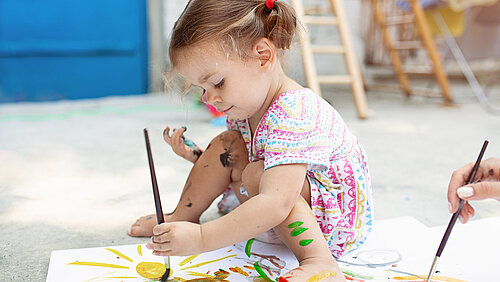 Kleines Mädchen sitzt auf dem Boden und malt mit Wasserfarben 