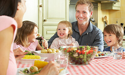 Familie_mit_drei_Kindern_sitzt_am_Essenstisch_mit_gesunden_Lebensmitteln