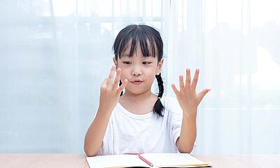 Kleines Mädchen sitzt vor einem Schulbuch und zaehlt mit den fingern