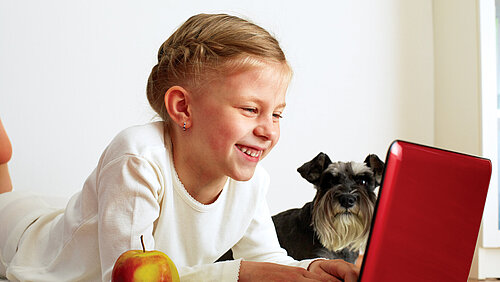 Junges Mädchen ist am Laptop und hinter ihr sitzt ein Hund