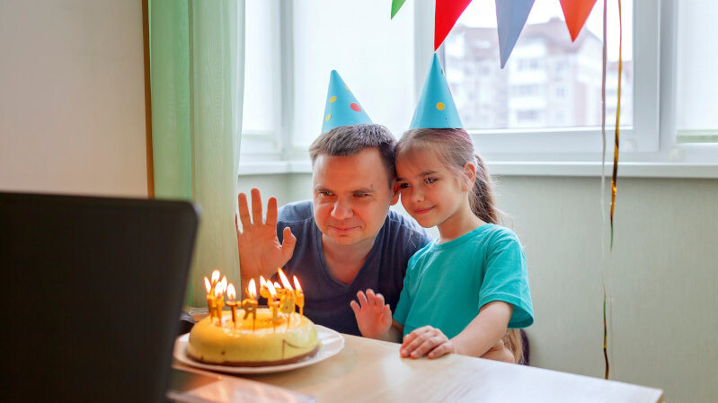 Vater_und_Tochter_stehen_mit_Geburtstagstorte_vor_Laptop_und_winken