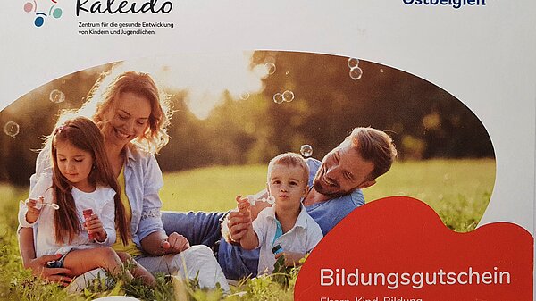 Familie sitzt auf einer Wiese und macht Seifenblasen. In der rechten unteren Ecke befindet sich ein Schriftzug mit dem Titel &quot;Bildungsgustchein&quot;.