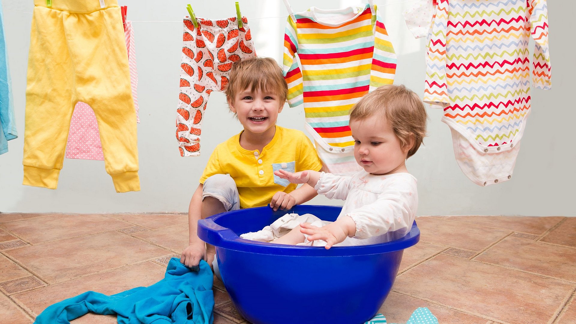 Bruder und Schwester spielen gemeinsam mit Kleidungsstücken. Die kleine Schwester sitzt im Wäschekorb.