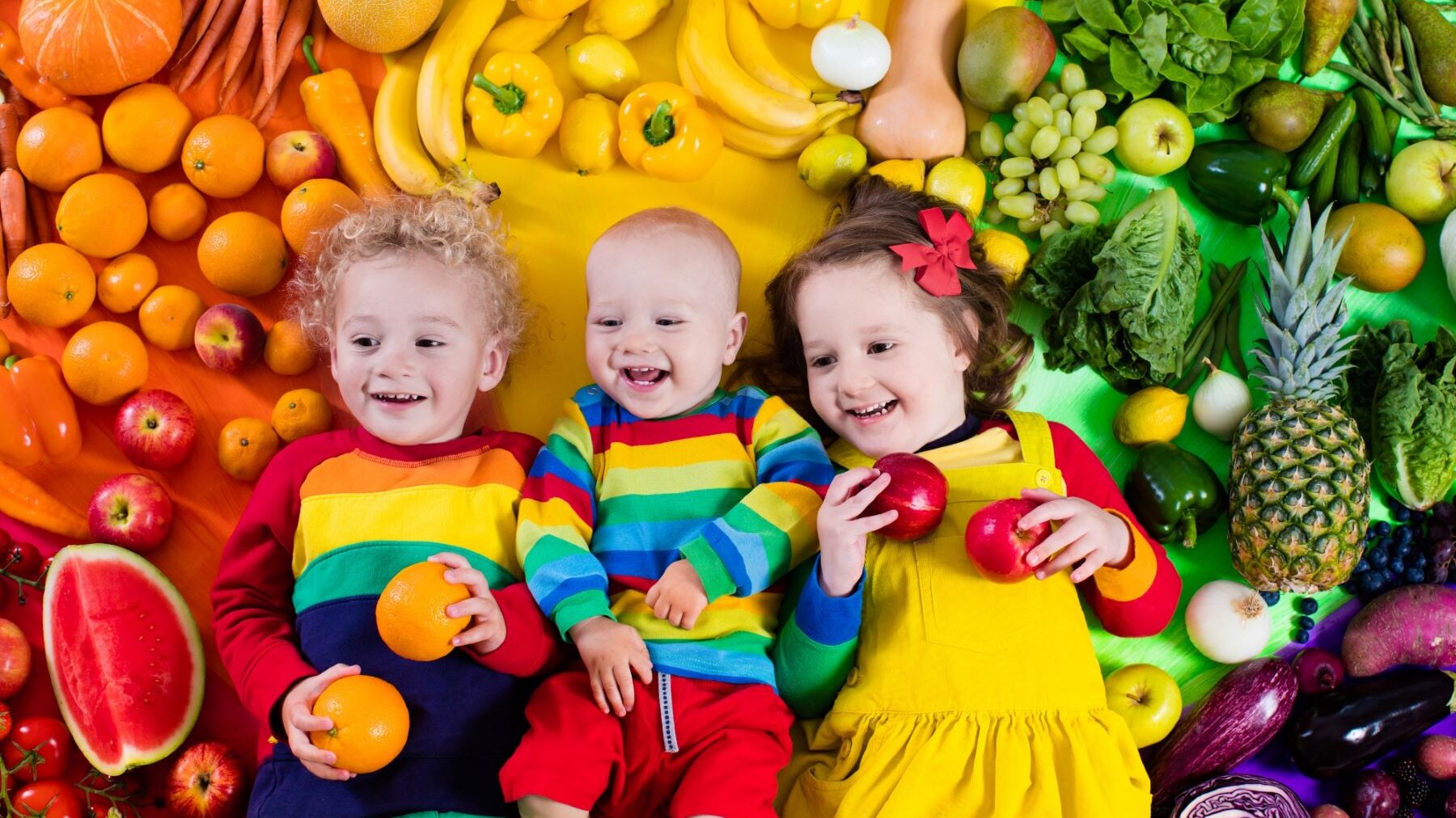 Kinder liegen zwischen buntem Obst und Gemüse, welches nach den Regenbogenfarben angerichtet ist. 