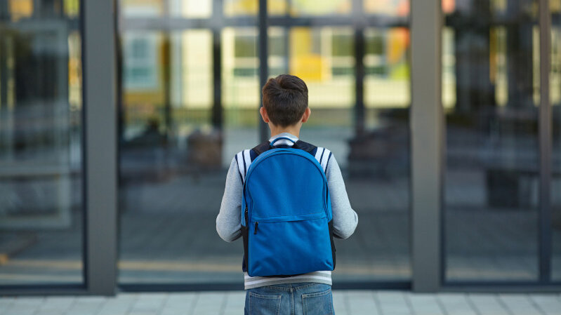 Kind steht mit Rucksack for den großen Glasstüren einer Schule