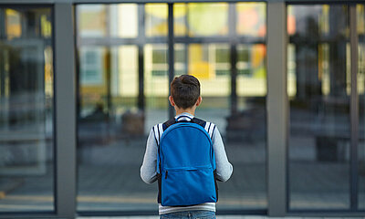 Kind steht mit Rucksack for den großen Glasstüren einer Schule